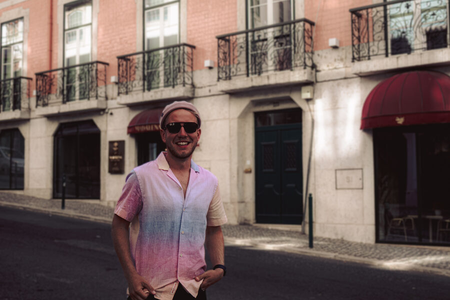 André smiling in a sunlit street, in sunglasses and a pastel shirt, hands in his pockets.