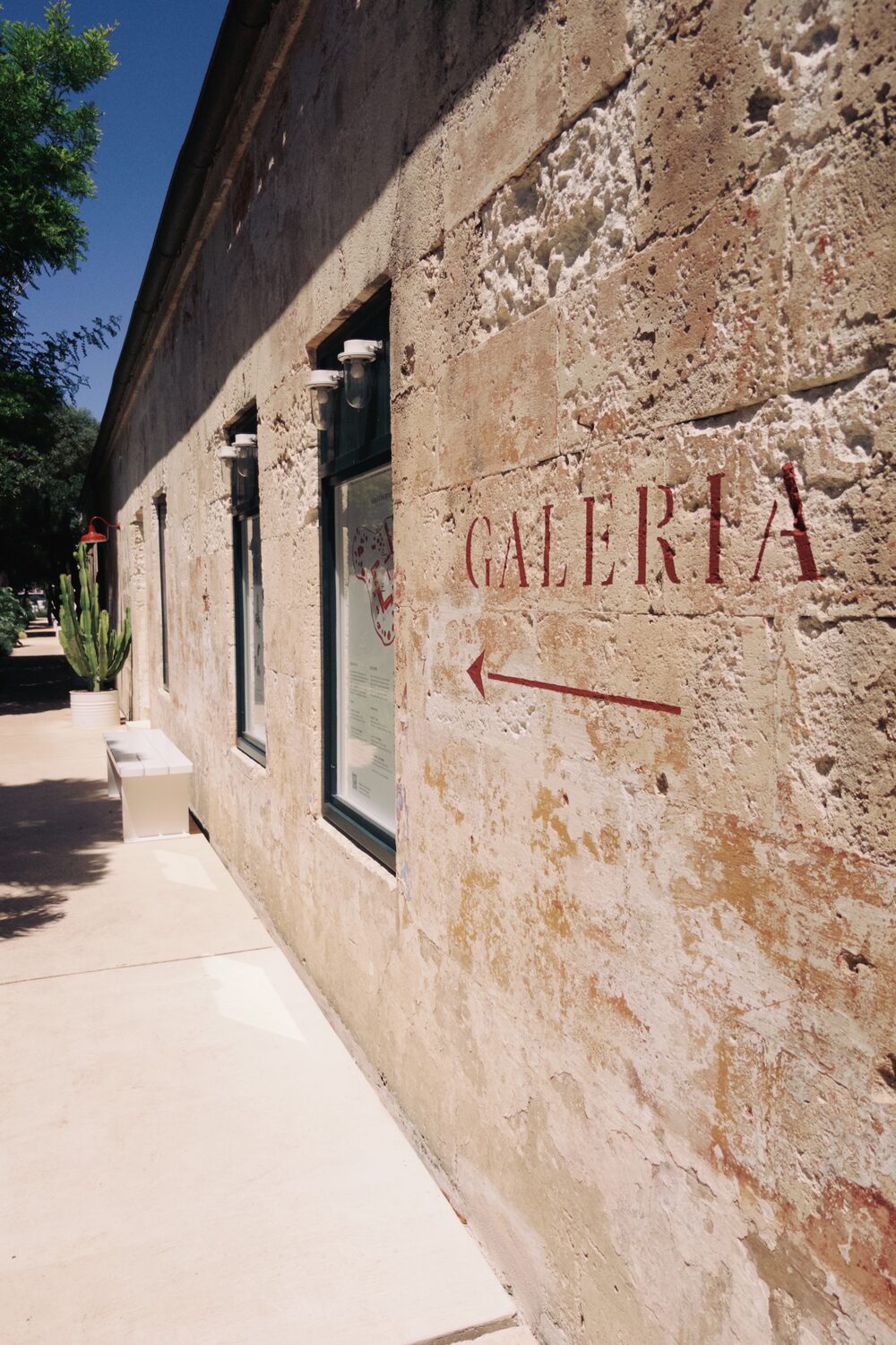 A weathered ochre adobe wall in bright sun, the word GALERÍA hand-painted in deep red beside an arrow pointing the way, past a window and a stone bench.